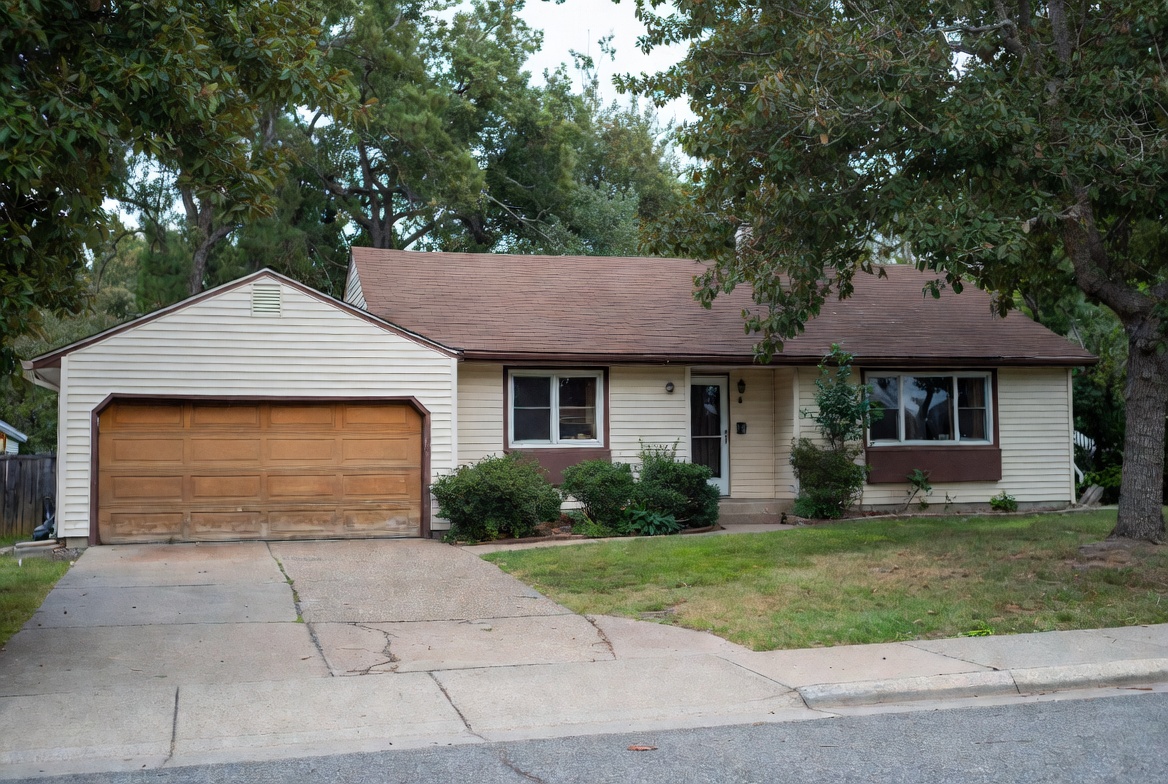 1980s ranch house with cream siding, brown trim, attached garage, driveway, lawn, and shrubs.