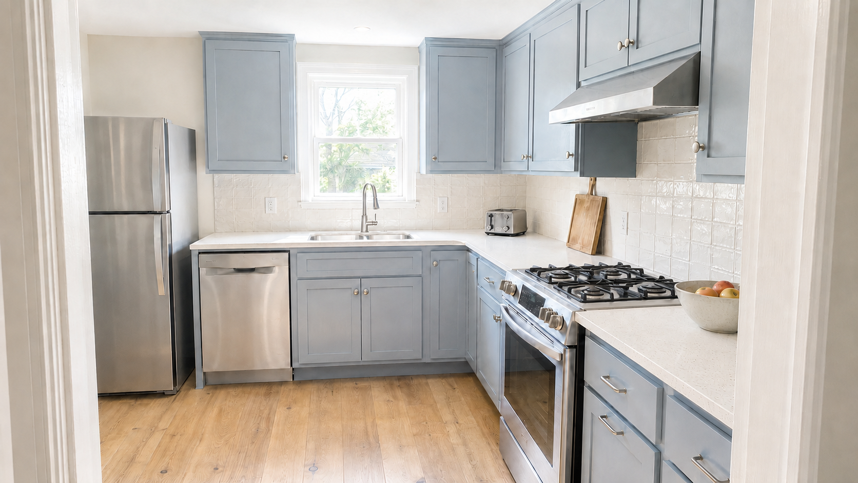 Muted blue-gray cabinets in a bright U-shaped kitchen with warm oak flooring.