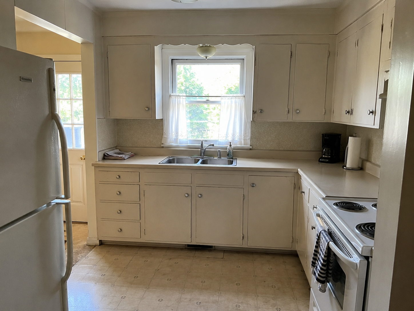 Older small Cape kitchen with sink window, worn cabinets, range, fridge, and tight layout.