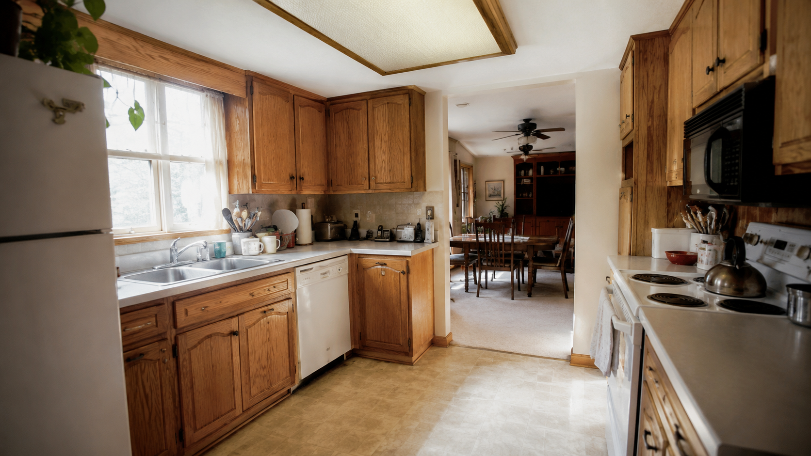 1980s ranch kitchen with oak cabinets, laminate counters, fluorescent ceiling light, and dining room beyond.