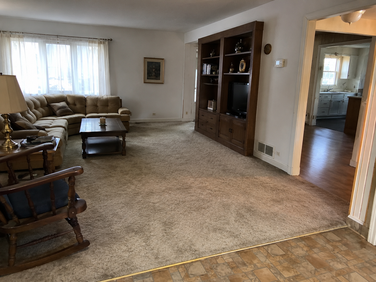 1980s ranch living room with beige carpet, flooring transition, low ceiling, and kitchen beyond.