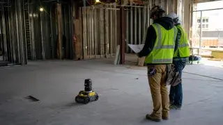 Construction workers reviewing plans while an AI-assisted layout robot marks a concrete floor inside an unfinished building.