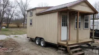 Budget tiny house on wheels with simple siding, metal roof, small porch, trailer frame, and gravel yard.