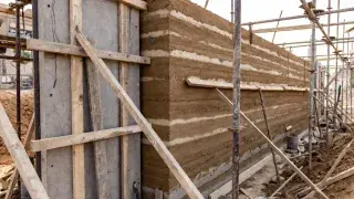 Rammed earth wall under construction with formwork, scaffolding, and visible layered compaction.