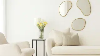Elegant minimalist living room with a white sofa, soft pillows, and gold-edged organic mirrors on a smooth warm white wall.