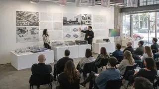 Architecture students presenting thesis boards and models to a seated review audience in studio.