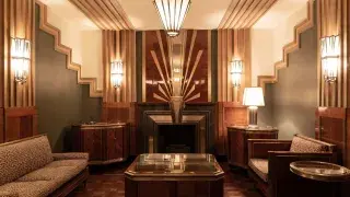 Art Deco sitting room with stepped wall geometry, polished wood paneling, symmetrical lighting, and a sunburst fireplace design.