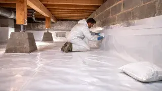 Worker sealing crawl space vapor barrier to the foundation wall with tape; vent cover visible on the left.