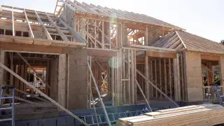 Wood-framed house under construction showing wall studs, partial sheathing, roof framing, bracing, and stacked lumber on site.
