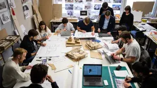 Architecture students working around a studio table with drawings, models, laptops, and project materials.