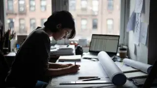 Entry-level architecture worker drawing at a desk near a New York City window.