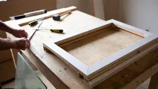Partly assembled wood window sash on a workbench with glass and tools nearby.