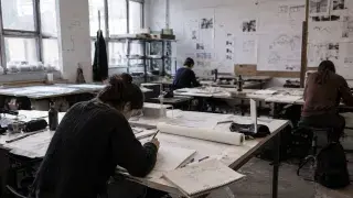 Architecture student working alone at a drafting table in a rough school design studio with drawings, models, and pinned project boards.