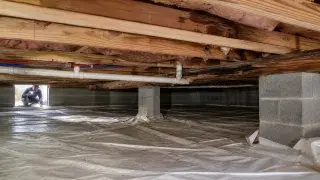 Crawl space inspection showing wood framing, insulation, pipes, block supports, and a ground vapor barrier.