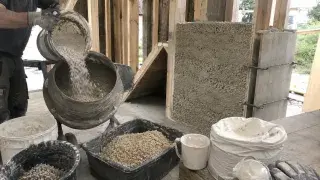 Worker pouring dry binder into a mixer beside hemp hurds, lime bags, buckets, timber framing, and a partially filled hempcrete wall on a small construction site.