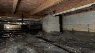 Low-clearance crawl space with ductwork, plumbing, block piers, and wrinkled ground liner that make inspection difficult.