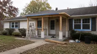 Ranch house front porch under construction with new posts, low roof, ladder, taped windows, tools, lumber, and an unfinished front yard.