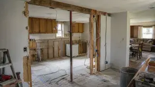 Ranch kitchen wall removal with temporary support, exposed studs, beam area, wiring, protected floor, and living room beyond.
