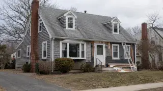 Older Cape Cod house exterior during remodeling with ladder, patched siding, dormers, and ordinary yard.