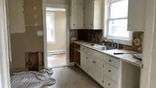 Small Cape Cod kitchen during remodeling with old cabinets, patched wall, window, and narrow layout.