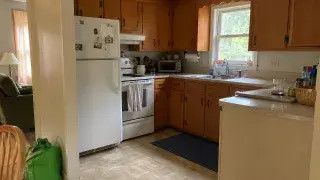 Dated Cape Cod kitchen with older wood cabinets, compact layout, white appliances, and modest finishes.