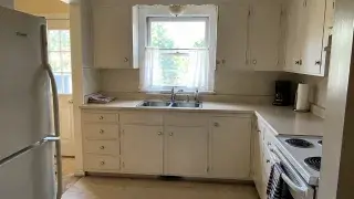 Older small Cape kitchen with sink window, worn cabinets, range, fridge, and tight layout.