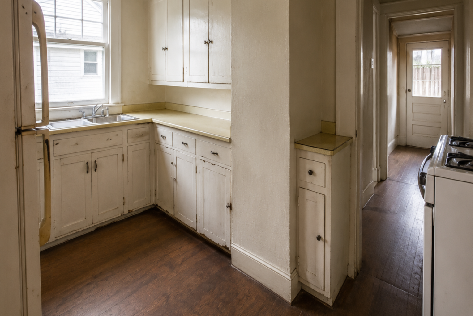 Unrenovated 1920s bungalow kitchen with worn cabinets, laminate counters, and a narrow layout.