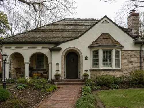 1940s-style stucco and stone cottage with arched entry, covered porch, bay window, chimney, weathered roof, and mature landscaping.