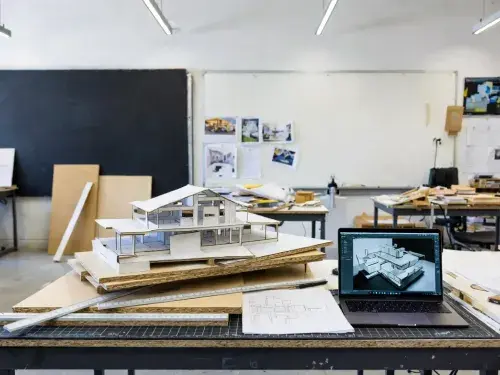 Architecture student studio workspace with physical massing models, drawings, and laptop.