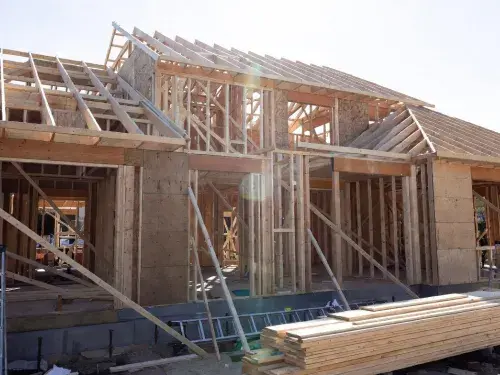 Wood-framed house under construction showing wall studs, partial sheathing, roof framing, bracing, and stacked lumber on site.