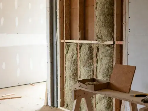 Careful mineral wool batt installation in open stud wall with cellulose and cork samples on workbench, premium construction detail.