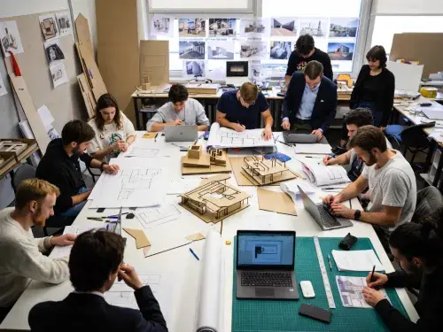 Architecture students working around a studio table with drawings, models, laptops, and project materials.