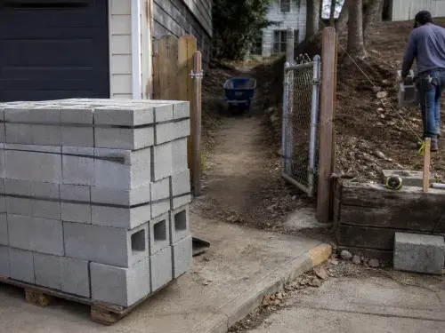 Concrete block pallet staged near a driveway with a narrow access path to the wall location.