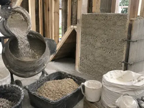 Worker pouring dry binder into a mixer beside hemp hurds, lime bags, buckets, timber framing, and a partially filled hempcrete wall on a small construction site.