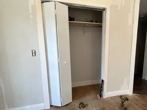 Partly installed white bifold closet door in an older home with the top track visible, tools on the floor, and a level and shim used to check the door opening.