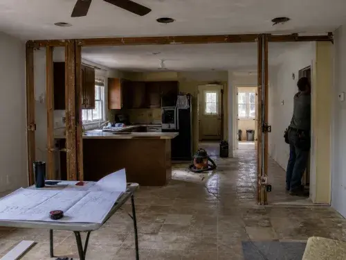 Small ranch house interior remodel with partial wall opening, exposed framing, low ceiling, old kitchen, and plans on a table.