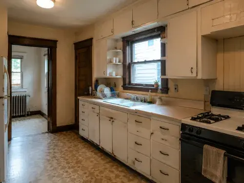 Old 1920s kitchen layout with sink under window, worn cabinets, stove, and radiator room beyond.
