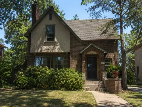 1930s brick and stucco house with steep gable, arched entry, mature shrubs, and front walkway.