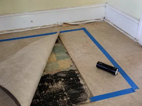 Lifted flooring in a 1950s house revealing old square vinyl tiles and black adhesive.