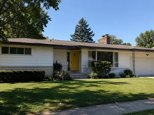 White 1950s ranch house with yellow front door, picture window, attached garage, and broad lawn.
