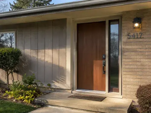 Updated 1970s front door with wood slab, sidelight, brick, modern hardware, and entry lighting.  Caption A good 1970s front door tells you more than the door style; the glass,