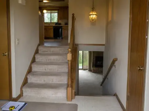 1980s split-level entry with stairs up and down, oak railing, beige carpet, brass light, and renovation notes.