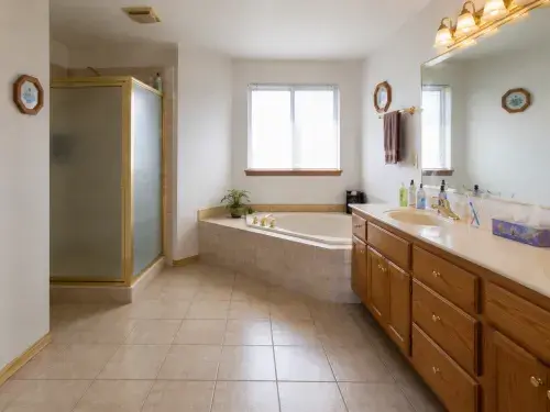 1990s primary bathroom with separate shower, deck tub, oak vanity, beige tile, and large mirror.