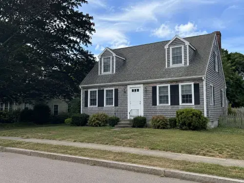 Documentary-style photo of a full Cape Cod house with cedar shakes, dormers, and a simple front facade.