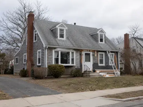 Older Cape Cod house exterior during remodeling with ladder, patched siding, dormers, and ordinary yard.