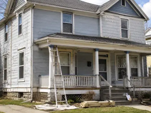 Early-1900s wood house in bright daylight with weathered siding, a covered porch, ladder, lumber and visible exterior repair planning.