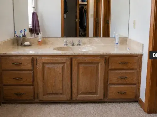 1980s bathroom vanity with oak cabinet, yellow cultured marble top, large mirror, dated faucet, and carpeted floor.