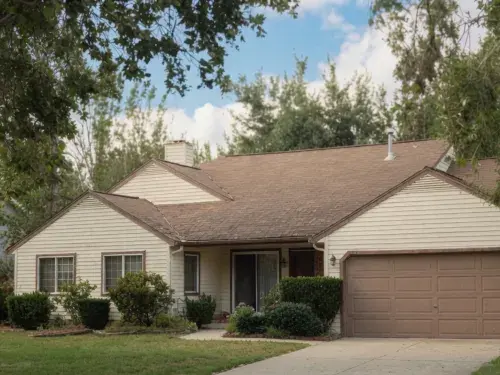 1980s suburban ranch house with cream siding, brown garage door, driveway, lawn, and shrubs.