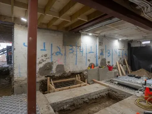 Basement underpinning and lowering project with staged excavation bays, soil removal access, exposed utilities, and new concrete support work.