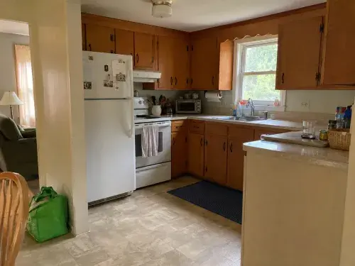 Dated Cape Cod kitchen with older wood cabinets, compact layout, white appliances, and modest finishes.